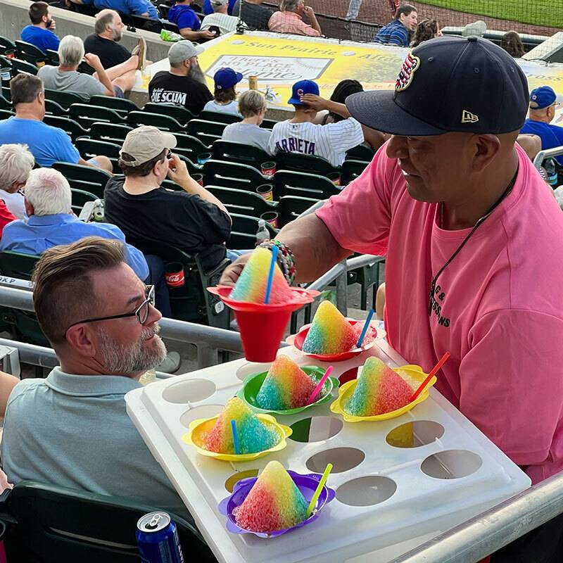 Man in pink t-shirt and navy ball cap standing in ballpark stadium seats holding a tray of sno-cones handing a sno-cone in a red cup to someone